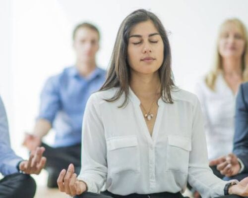 A woman practising wellness and mental health meditating in front of a group of business people.